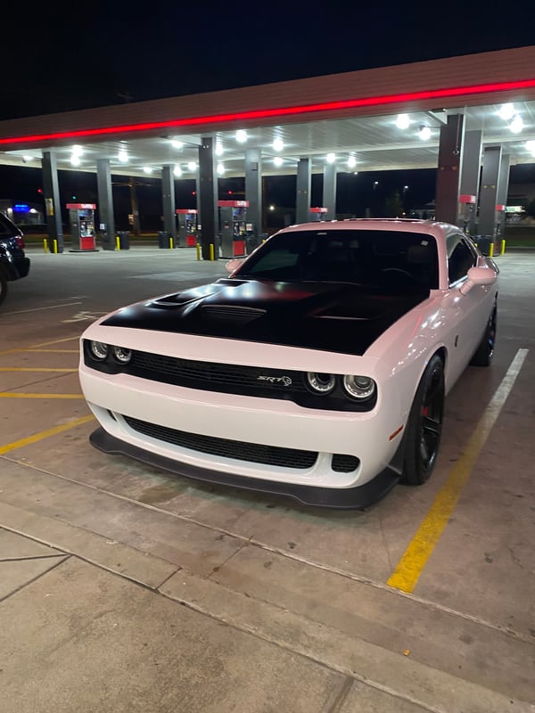 A phone photo of a white Dodge Challenger Hellcat at a gas station at night